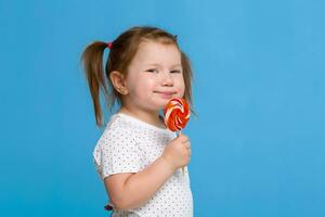 Beautiful little female child holding huge lollipop spiral candy smiling happy isolated on blue background. photo