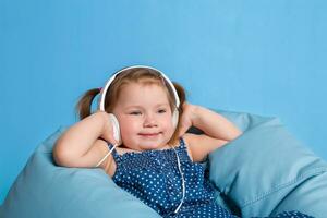 linda pequeño niña en auriculares escuchando a música utilizando un tableta y sonriente mientras sentado en azul grande bolso foto