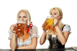 Two beautiful women drink beer from large glasses while sitting at a wooden table on a white background in the studio photo