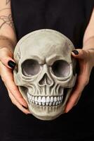 Tattooed hands of a woman in a black watch and clothes are holding a realistic model of a human skull with teeth. Close-up shot. photo