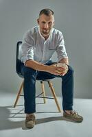 Man with stylish mustache, in classic white shirt and blue trousers is sitting on chair, holding glass of whiskey. Grey background, close-up shot. photo