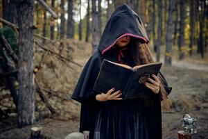 Witch in black dress with cape and hood. Holding book, pronouncing spell, posing in pine forest at table with accessories for witchcraft. Close-up. photo