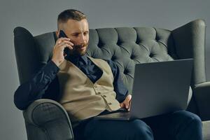 Man with stylish mustache, dressed in black shirt and beige vest is sitting on dark sofa, using his laptop and smartphone. Grey background, close-up. photo