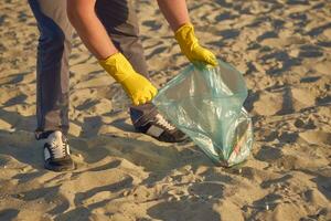Young volunteer in yellow gloves is walking with garbage bag along a dirty beach of the river and cleaning up trash. People and ecology. Close-up. photo