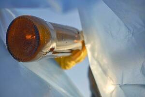 Hand of a conscious person in a yellow rubber glove puts a used brown glass bottle in a blue trash bag. Blue sky. Riverside pollution. Close-up shot. photo