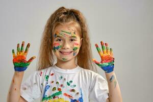 Little girl in white t-shirt is posing standing isolated on white and gesticulating with her painted in different colors palms. Art studio. Close-up. photo