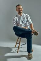 Man with stylish mustache, in classic white shirt and blue trousers is posing sitting on chair. Grey background, close-up shot. photo