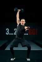 Strong brutal man doing squats with dumbbell at gym, full length photo. Guy doing exercises with dumbbell indoors. photo