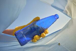 Hand of a conscious person in a yellow rubber glove puts a used blue glass bottle in a trash bag. Blue sky. Riverside pollution. Close-up shot. photo