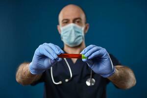 Doctor in mask, blue gloves, medical suit. Holding red laboratory test tube with green cap in hands. Blue background. Coronavirus, COVID-19. Close up photo