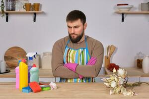 joven hermoso barbado hombre en el cocina, muestra todas su limpieza personal - detergentes, cepillos, aerosoles él pensar él es Listo para real limpieza foto