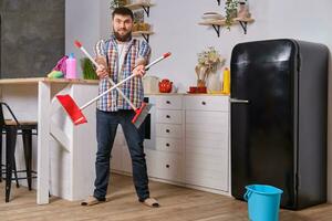 Young handsome bearded man in the kitchen, wearing checkered shirt, tries to deal with a dustpan and a broom. photo