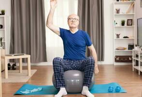 Pensioner doing exercise at home using swiss ball. Old person pensioner online internet exercise training at home sport activity with dumbbell, resistance band, swiss ball at elderly retirement age. photo