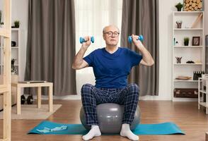 Senior man with vitality exercising in living room. Old person pensioner online internet exercise training at home sport activity with dumbbell, resistance band, swiss ball at elderly retirement age. photo