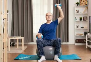 Grandfather using dumbbells being careful with his health. Old person pensioner online internet exercise training at home sport activity with dumbbell, resistance band, swiss ball at elderly retirement age. photo