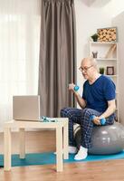 Senior man training biceps watching online fitness lesson. Old person pensioner online internet exercise training at home sport activity with dumbbell, resistance band, swiss ball at elderly retirement age. photo