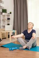 Retired woman relaxing in the living room on yoga mat Old person pensioner online internet exercise training at home sport activity with dumbbell, resistance band, swiss ball at elderly retirement age photo