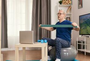 Strong old man training online with resistance band. Old person pensioner online internet exercise training at home sport activity with dumbbell, resistance band, swiss ball at elderly retirement age. photo