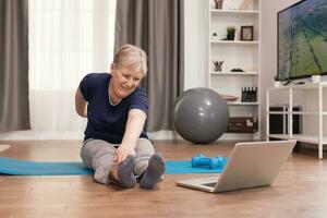Cheerful old woman doing sports watching online lessons. Old person pensioner online internet exercise training at home sport activity with dumbbell, resistance band, swiss ball at elderly retirement age photo