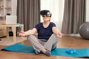 Grandmother wearing VR headset sitting on yoga mat. Old person pensioner online internet exercise training at home sport activity with dumbbell, resistance band, swiss ball and VR headset at elderly retirement age photo