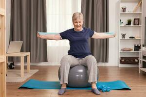 Senior woman working out at home with fitness bands to solve her health problems. Old person pensioner online internet exercise training at home sport activity with dumbbell, resistance band, swiss ball at elderly retirement age photo