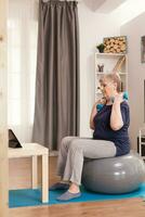 Old woman lifting dumbbells sitting on stability ball watching training lesson on laptop. Old person pensioner online internet exercise training at home sport activity with resistance band, swiss ball at elderly retirement age photo