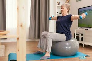 Old housewife using dumbbells for physical recovery at home. Old person pensioner online internet exercise training at home sport activity with resistance band, swiss ball at elderly retirement age photo