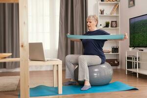 Elderly woman workout at home in front of the laptop. Old person pensioner online internet exercise training at home sport activity with dumbbell, resistance band, swiss ball at elderly retirement age photo