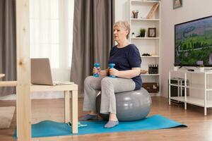 Old housewife watching online fitness lesson at home. Old person pensioner online internet exercise training at home sport activity with dumbbell, resistance band, swiss ball at elderly retirement age photo