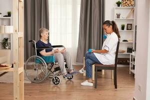 Doctor showing to immobilized old woman exercises with dumbbells for physical recovery. Disabled handicapped old person recovering professional help nurse, nursing retirement home treatment and rehabilitation photo