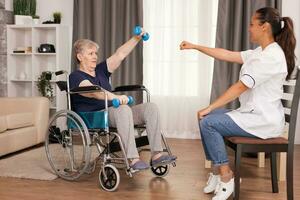 Invalid old woman training her arms using dumbbells. Disabled handicapped old person recovering professional help nurse, nursing retirement home treatment and rehabilitation photo