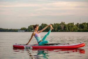 Young woman sitting on paddle board, practicing yoga pose. Doing yoga exercise on sup board, active summer rest photo