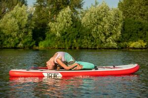 Young woman doing yoga on sup board with paddle. Yoga pose, side view - concept of harmony with the nature. photo
