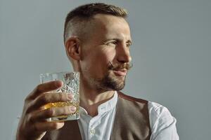 Man with stylish mustache, dressed in classic brown vest, white shirt is sitting at the table, enjoying whiskey. Grey background, close-up shot. photo