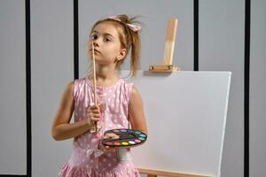 Little girl artist in a pink dress is standing behind easel and painting with brush on canvas at art studio with white walls. Medium close-up shot. photo