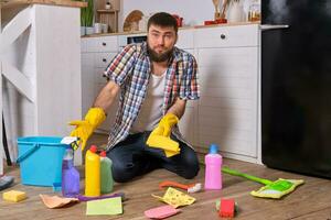 Caucasian young bearded man sits on the floor of his kitchen and tries to clean it using all of his detergents, rags, washcloths and brushes photo