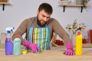 joven hermoso barbado hombre en el cocina, molesto a limpiar un mesa utilizando detergentes, cepillos, aerosoles foto