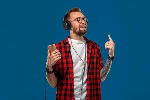 hermoso joven elegante hombre en auriculares participación mp3 jugador y bailando mientras en pie en contra azul antecedentes foto