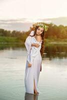 Beautiful black haired girl in white vintage dress and wreath of flowers standing in water of lake. Sun flare. photo