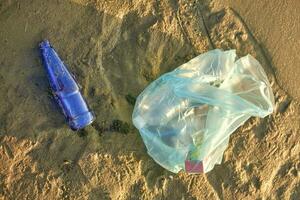 Used blue glass bottle and garbage bag lies on the river beach. Cleaning up trash. Riverside pollution. Volunteering concept. Close-up shot. photo