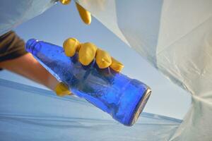 mano de un consciente persona en un amarillo caucho guante pone un usado azul vaso botella en un basura bolsa. azul cielo. orilla contaminación. de cerca disparo. foto