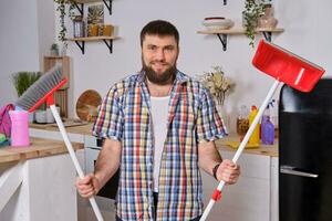 Young handsome bearded man in the kitchen, wearing checkered shirt, tries to deal with a dustpan and a broom. photo