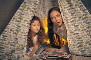 Mother and daughter are sitting in a teepee tent with some pillows and reading stories. Happy family. photo