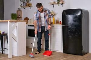 Young handsome bearded man in a kitchen, wearing a plaid shirt, sweeping the floor with a shovel and broom. photo