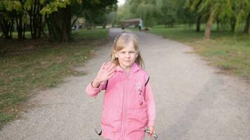 a little girl in a pink jacket walks along a walking path in the park and waves hello or bye video