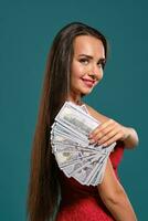 Brunette girl with a long hair, wearing a sexy red dress is posing holding a fan of hundred dollar bills against a blue background. photo