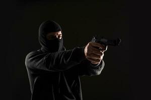 The robber in a mask with a gun pointed to the side on a black background photo