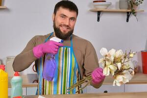 joven hermoso barbado hombre en el cocina, muestra todas su limpieza personal - detergentes, cepillos, aerosoles él pensar él es Listo para real limpieza foto