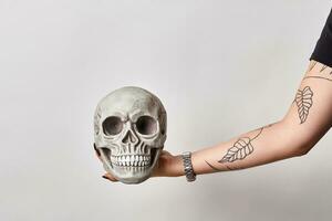 Tattooed hand of a woman in a black watch is holding a realistic model of a human skull with teeth isolated on white. Close-up shot. photo