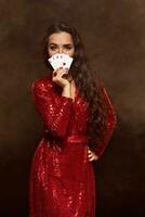 Young beautiful lucky brunette in a red evening dress shows her cards, a four of aces, hiding her face with cards photo